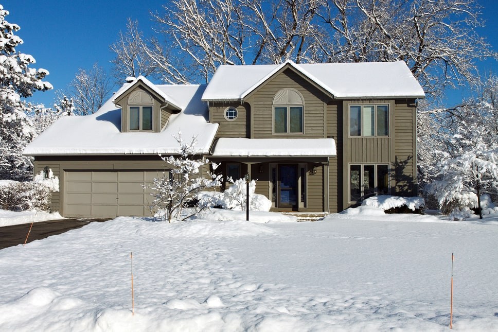 A brown siding home with several inches of snow piled on the roof and in the front yard.