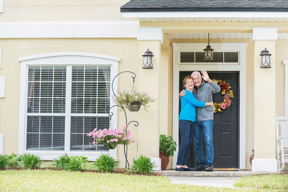 A married couple smiling and hugging in front of their older home they have recently renovated.